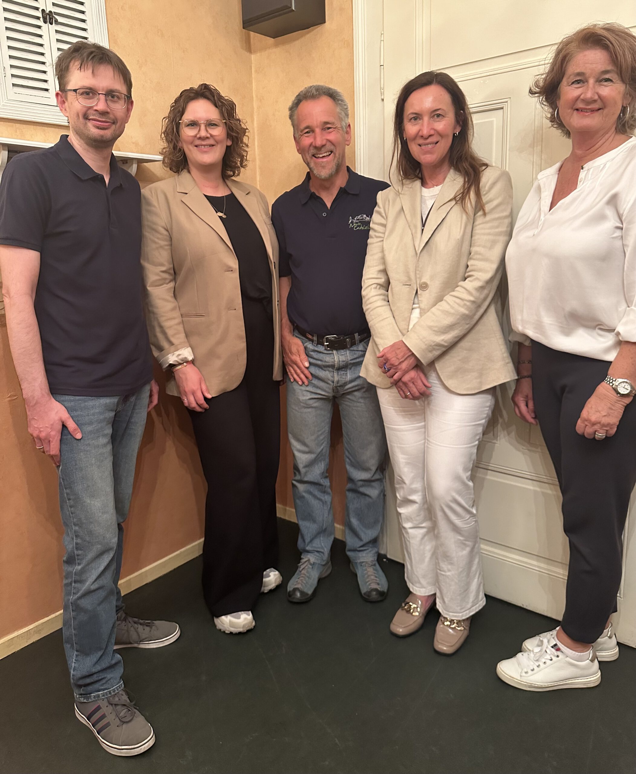 Das Foto zeigt den neu gewählten Vorstand der Grünen in Cadolzburg, posierend in einem Raum mit warm getönten Wänden. Von links nach rechts stehen Patrick Carl in einem dunklen T-Shirt und Jeans, Lisa Gernbacher mit lockigem Haar, gekleidet in einen dunklen Blazer über einer Bluse, Dieter Burock in einem blauen Poloshirt und Jeans, Sabine Geyer in einem hellen Jackett und weißen Hosen, und Uschi Schröder in einer weißen Bluse und einer hellen Hose. Alle Mitglieder lächeln und blicken direkt in die Kamera, was ein Gefühl von Zusammenhalt und Engagement vermittelt.