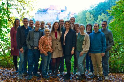 Eine Gruppe von sechzehn Erwachsenen steht in zwei Reihen im Freien und blickt in die Kamera. Sie tragen gemischte legere und formelle Kleidung wie Blazer, Cardigans, Jeans, Stiefel und eine beige Kostümjacke. Der Boden ist mit Herbstlaub bedeckt, seitlich stehen grüne Büsche und Bäume. Im Hintergrund ist die Cadolzburg deutlich zu erkennen. Kontext: Gruppenfoto der Kandidierenden der Grünen in Cadolzburg zur Gemeinderatswahl.