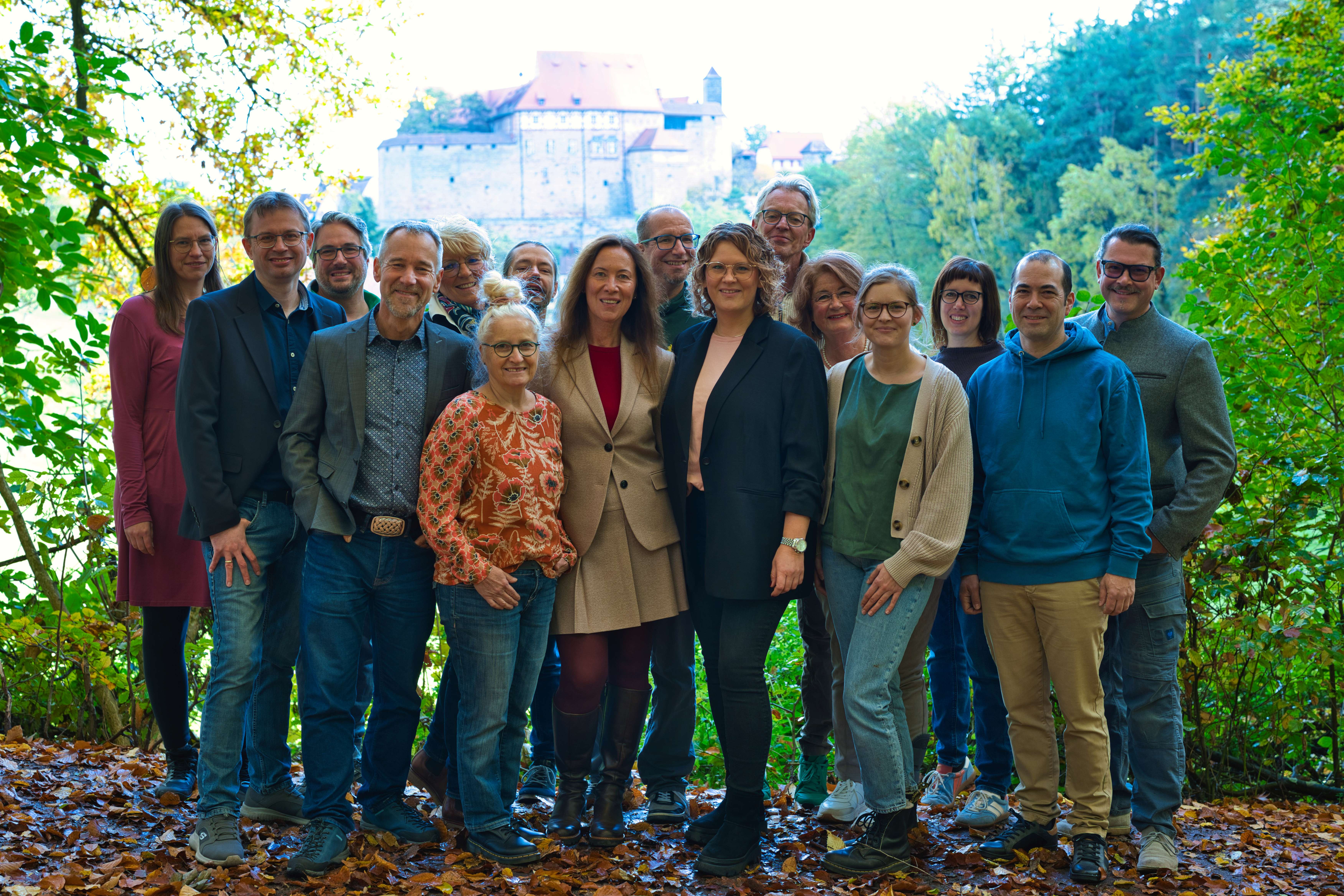 Eine Gruppe von sechzehn Erwachsenen steht in zwei Reihen im Freien und blickt in die Kamera. Sie tragen gemischte legere und formelle Kleidung wie Blazer, Cardigans, Jeans, Stiefel und eine beige Kostümjacke. Der Boden ist mit Herbstlaub bedeckt, seitlich stehen grüne Büsche und Bäume. Im Hintergrund ist die Cadolzburg deutlich zu erkennen. Kontext: Gruppenfoto der Kandidierenden der Grünen in Cadolzburg zur Gemeinderatswahl.